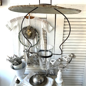 Puede incluir: Una exhibición de estilo antiguo con una estructura metálica colgante, un portavasos y candelabros. Un vestido blanco cuelga en el fondo, junto con un jarrón blanco, una pequeña planta y una mantequera plateada. El conjunto es vintage y decorativo.