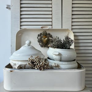 Può includere: Un portapane in metallo color crema contenente due zuppiere in ceramica bianca con coperchi e manici decorati. Una zuppiera contiene un mazzetto di lavanda essiccata, mentre davanti si trova un grappolo di bacche bianche essiccate. Un cuore sacro in metallo è appeso all'interno.