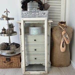 Peut inclure: Une présentation de style vintage avec un présentoir à étages, une armoire blanche à tiroirs et un mannequin de couture. Une couronne de baies noires et une boîte décorative sont également présentes. Des chaussons de ballet sont suspendus au mannequin.
