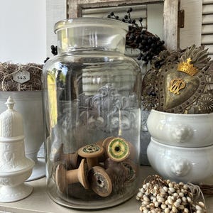 Op de afbeelding: Een grote, doorzichtige glazen apothekerspot met een cilindrisch lichaam en een plat deksel, gevuld met diverse vintage houten garenklosjes. De klosjes zijn voorzien van ronde papieren etiketten op de uiteinden met gedrukte tekst.