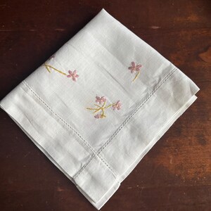 May include: A folded white linen napkin with embroidered pink flowers and yellow stems. The napkin has a stitched border and is set against a dark wood surface. The floral embroidery adds a delicate touch to the classic design.