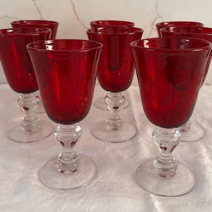 May include: A set of eight ruby red wine glasses. Each glass features a clear, decorative stem and a wide, rounded bowl. The glasses are arranged on a white surface, showcasing their vibrant color and elegant design.