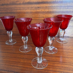 May include: Five red glass goblets with clear stems and bases are arranged on a wooden surface. The goblets have a tulip-shaped bowl and a clear, decorative stem. The red glass has a glossy finish.