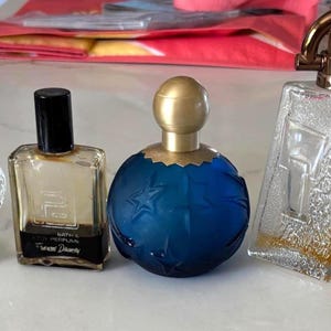 May include: Five vintage perfume bottles in different shapes and sizes. The bottles are clear, blue, and black, with gold and black detailing. One bottle is crown-shaped, and another has a blue globe design. The bottles are arranged in a line.