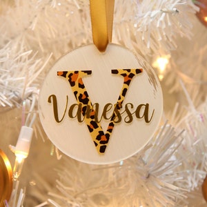 Peut inclure: Ornement de Noël rond blanc avec un ruban doré. L'ornement a une lettre dorée "V" avec un motif léopard et le nom "Vanessa" écrit en lettres dorées.