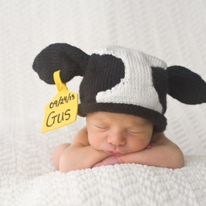 May include: A newborn baby wearing a black and white cow hat with large ears and a yellow tag that reads "09/24/13" and "Gus". The baby is lying on a white blanket with their eyes closed.