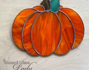 May include: A stained glass pumpkin suncatcher featuring vibrant orange swirled glass panels outlined in dark metal solder. The pumpkin has two distinct green glass leaves at the top and a small metal loop for hanging, with the text Stained Glass Lady.