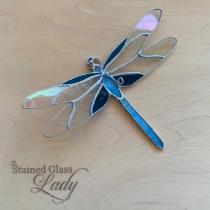 May include: A stained glass dragonfly with iridescent wings and a blue body. The dragonfly's wings are a mix of clear, light brown, and dark blue glass, with a silver metal outline. The text "The Stained Glass Lady" is in the lower left corner.