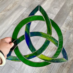 May include: A stained glass Celtic knot in shades of green and blue. The triangular design features interwoven lines, creating a three-part knot. The glass has a textured appearance, with a silver metal border.