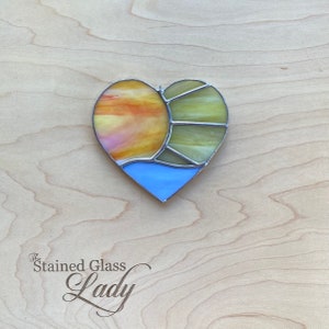 Puede incluir: Un atrapasol de vidrio coloreado en forma de corazón con un sol amarillo, naranja y rosa y una ola azul. El atrapasol está sobre un fondo de madera clara.