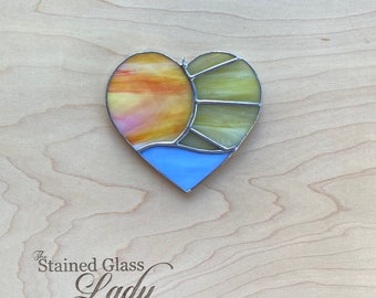 Mini enfeite de vitral artesanal em formato de coração, representando um pôr do sol em tons de azul claro, amarelo e amarelo-alaranjado. Uma ótima ideia de presente com temática praiana, perfeito para decorar sua casa ou carro.