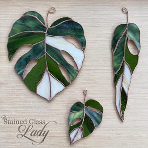 Vitral artesanal em formato de folha de monstera nas cores verde-samambaia e marfim, perfeito para pendurar na janela. Decoração natural para casa.