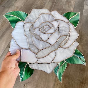 May include: A white stained glass rose with green leaves. The rose is made of many small pieces of glass that are joined together with lead. The rose is a popular motif in stained glass art.