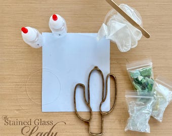Pode incluir: Um kit de artesanato contendo uma moldura de madeira em forma de cacto, dois pequenos frascos brancos com tampas vermelhas, um palito de madeira para misturar, um recipiente de plástico com luvas brancas e três sacos com pedaços de vidro verde e claro.