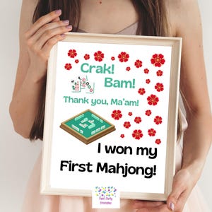 Pode incluir: Uma impressão emoldurada com o texto "Crak! Bam! Thank you, Ma'am! I won my First Mahjong!" rodeada por ilustrações de flores vermelhas e um tabuleiro verde de Mahjong. A impressão é segurada por uma pessoa.