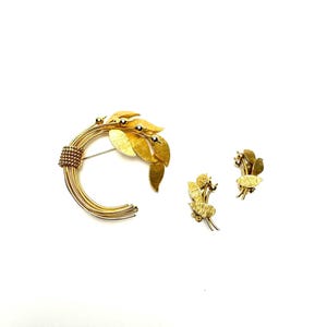 Könnte beinhalten: Ein goldfarbenes Schmuckset mit einer Brosche und Clip-Ohrringen. Die Brosche hat ein halbkreisförmiges Design mit einem Blatt- und Beerenmotiv. Die Ohrringe und die Brosche haben eine strukturierte Oberfläche auf weißem Hintergrund.