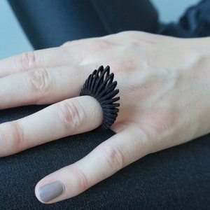 Puede incluir: Un anillo negro y puntiagudo se lleva en un dedo. El anillo tiene un diseño geométrico único. Las uñas de la persona están pintadas de un color gris neutro. El fondo es azul y negro suave y apagado.