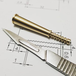 May include: A close-up of a brass-colored, conical object with a threaded base and a stainless steel scalpel. The objects are on a white surface with black lines and numbers, likely a technical drawing.