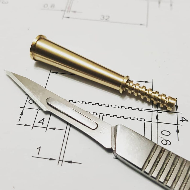 May include: A close-up of a brass-colored, conical object with a threaded base and a stainless steel scalpel. The objects are on a white surface with black lines and numbers, likely a technical drawing.