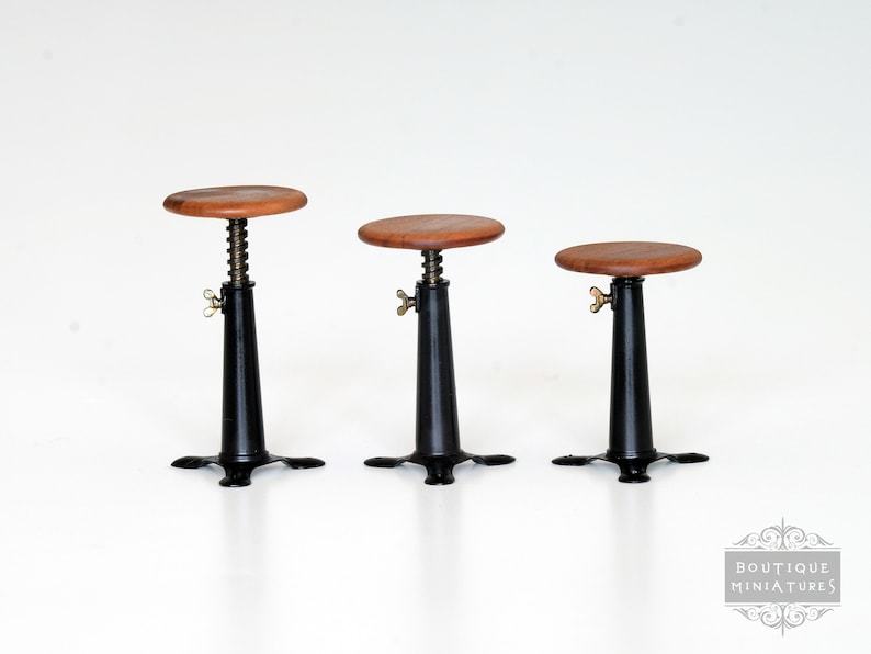 May include: Three miniature adjustable stools. Each stool has a round, wooden seat, a black metal base, and a brass-colored adjustment knob. The stools are designed to be adjustable in height.