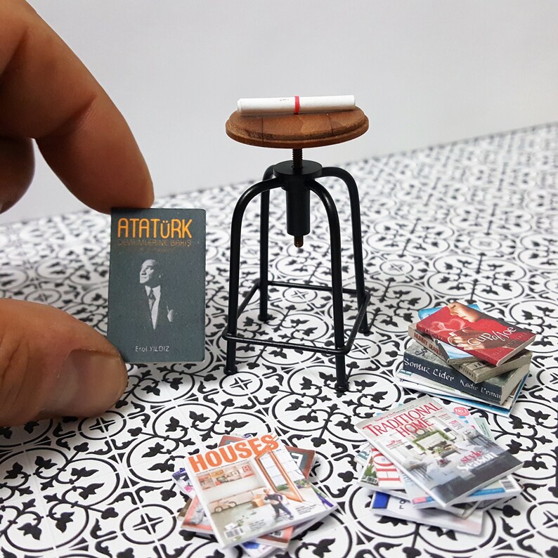 May include: A miniature black metal stool with a wooden seat and a rolled up piece of paper on top. The stool is surrounded by miniature books and magazines, including one with the title "ATAT&Uuml;RK" and another with the title "TRADITIONAL HOME".