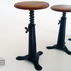 May include: Miniature black adjustable stools with wooden seats. The stools have a black base with three legs, a black stem with a metal spring, and a wooden seat. The stools are designed for a dollhouse or miniature setting.