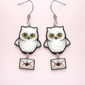 Peut inclure: Boucles d'oreilles en forme de hibou. Chaque boucle d'oreille représente un hibou blanc aux yeux jaunes et un petit charme en forme d'enveloppe avec un sceau de cire rouge. Les boucles d'oreilles sont suspendues à des crochets argentés.