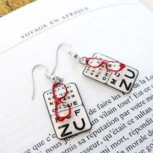 以下が含まれることがあります： シルバーカラーのドロップピアス。各イヤリングには、赤いフレームのメガネが付いた、小さな長方形の視力検査表があります。視力検査表には、黒い文字と数字が表示されています。