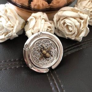 May include: A silver-toned metal purse hook with a bee design. The bee is made of gold-toned metal and is centered on the hook. The hook is round and has a decorative design around the edge.