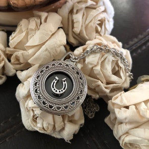 May include: A silver necklace with a round pendant featuring a black horseshoe charm. The pendant is surrounded by an intricate silver design. The necklace is lying on a surface with white flowers.