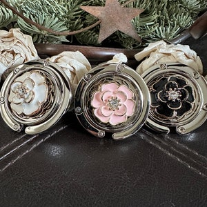 May include: Three silver-toned purse hooks with a flower design. The flowers are white, pink, and black with a rhinestone center.