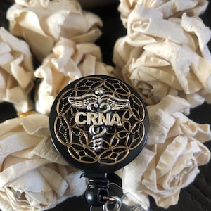 K&ouml;nnte beinhalten: Schwarzer und goldener ausziehbarer Badgehalter mit einem silbernen medizinischen Symbol und dem Text "CRNA".