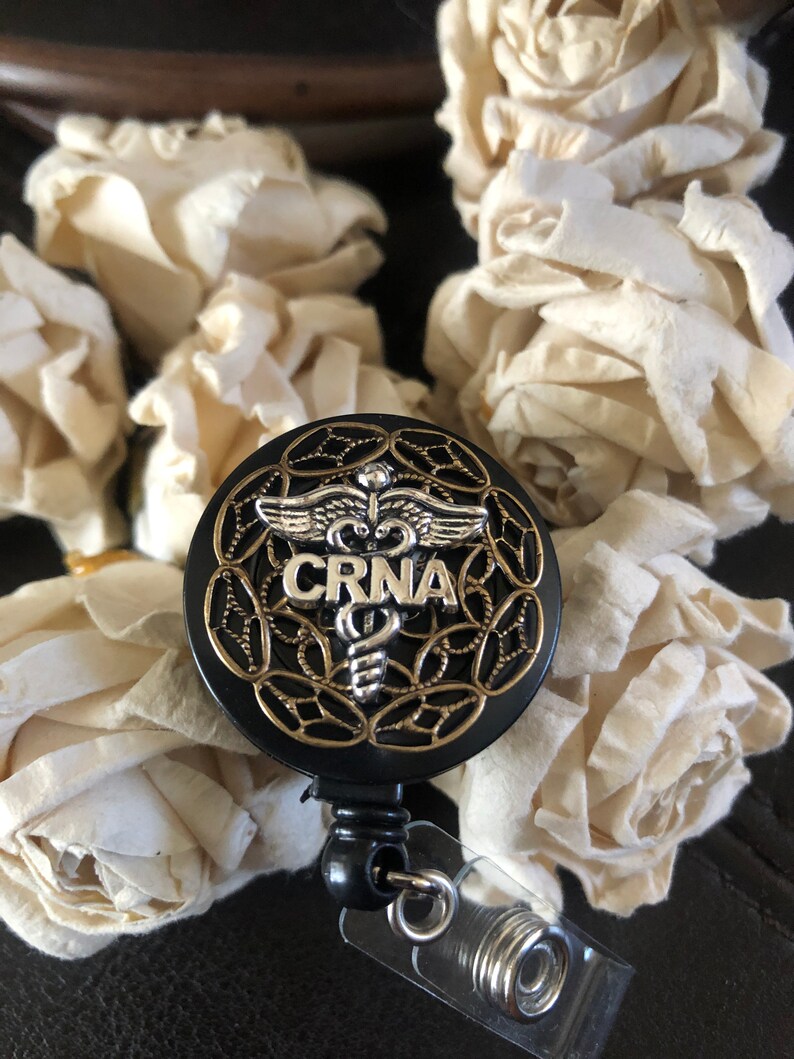 K&ouml;nnte beinhalten: Schwarzer und goldener ausziehbarer Badgehalter mit einem silbernen medizinischen Symbol und dem Text "CRNA".