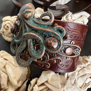 May include: A brown leather bracelet with an ornate copper and green octopus design. The octopus has a detailed texture and is set against a background of intricate floral patterns.