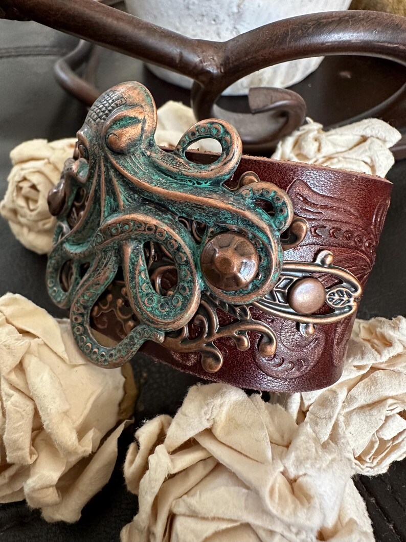 May include: A brown leather bracelet with an ornate copper and green octopus design. The octopus has a detailed texture and is set against a background of intricate floral patterns.