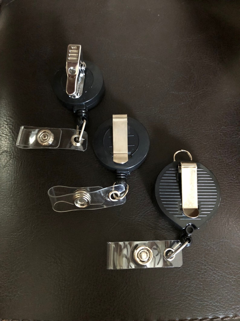 K&ouml;nnte beinhalten: Drei schwarze, einziehbare Ausweishalter mit durchsichtigen Plastikb&auml;ndern. Jeder Halter hat einen silbernen Clip und einen Knopf am Band.