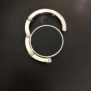 May include: A circular, foldable bag hanger featuring a polished silver-tone metal frame and a solid black center disc. The device consists of a hinged hook mechanism that unfolds from the main circular base, designed for suspending items.