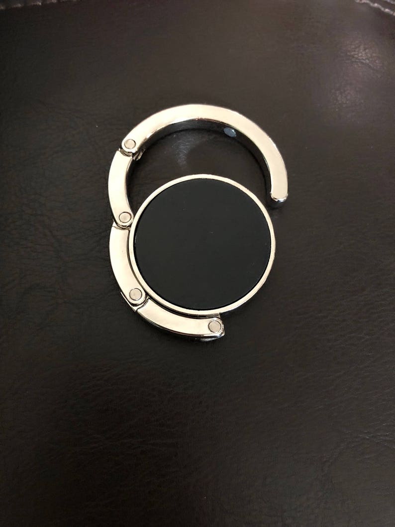 May include: A circular, foldable bag hanger featuring a polished silver-tone metal frame and a solid black center disc. The device consists of a hinged hook mechanism that unfolds from the main circular base, designed for suspending items.