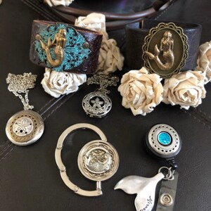 May include: A collection of mermaid-themed jewelry and accessories, including two leather bracelets with mermaid charms, a silver chain necklace with a seashell pendant, a silver retractable badge holder with a mermaid tail charm, and a silver key chain with a mermaid charm.