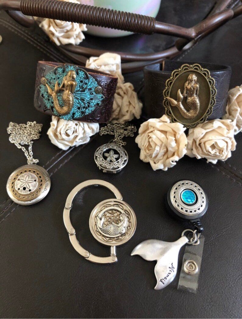 May include: A collection of mermaid-themed jewelry and accessories, including two leather bracelets with mermaid charms, a silver chain necklace with a seashell pendant, a silver retractable badge holder with a mermaid tail charm, and a silver key chain with a mermaid charm.
