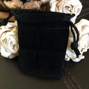 May include: A small, rectangular black velvet drawstring pouch featuring a soft, matte fabric texture and matching black braided cords on both sides. The pouch is designed with a simple top closure mechanism that cinches shut using the integrated cords.