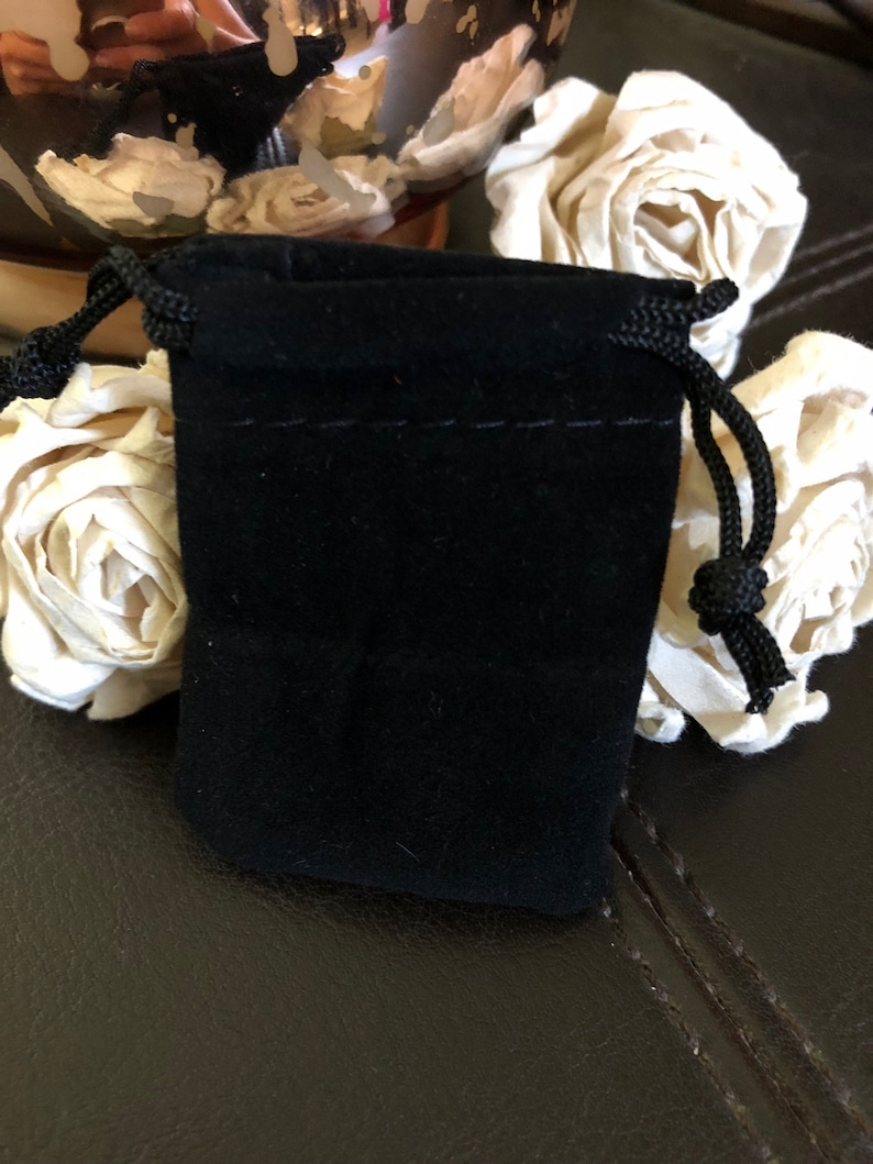 May include: A small, rectangular black velvet drawstring pouch featuring a soft, matte fabric texture and matching black braided cords on both sides. The pouch is designed with a simple top closure mechanism that cinches shut using the integrated cords.
