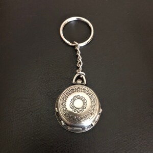 May include: A silver oval-shaped keychain with an intricate design and a small circle in the center. The keychain has a silver chain and a silver ring.