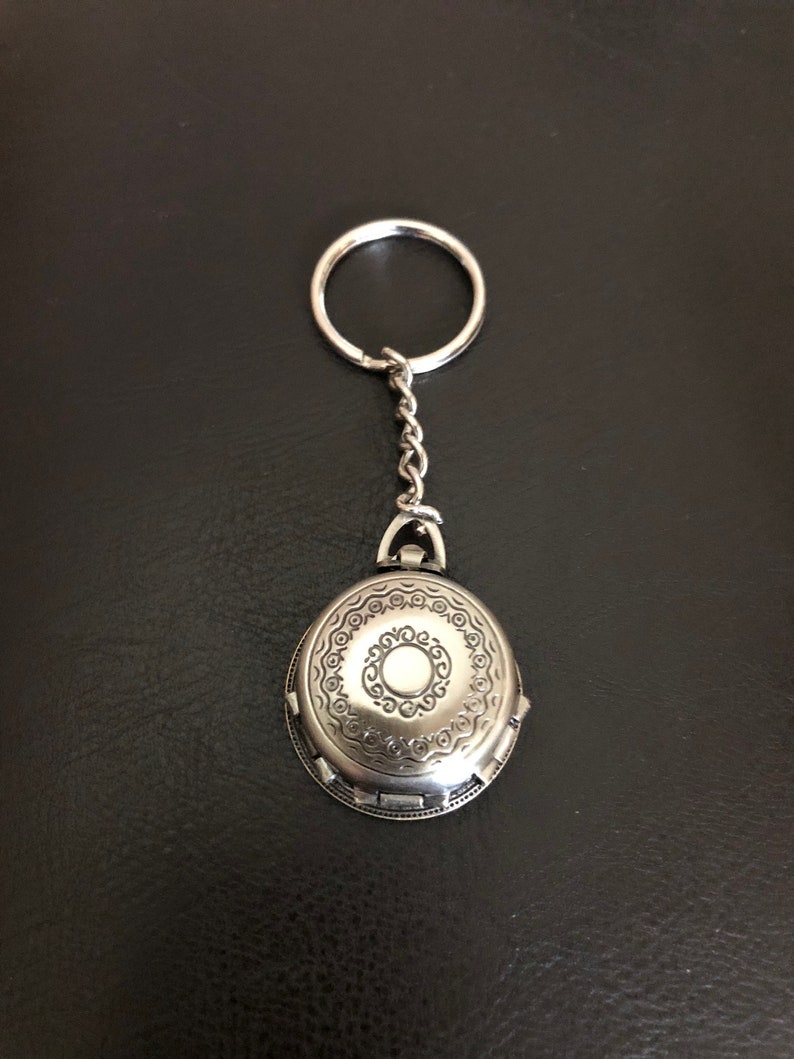May include: A silver oval-shaped keychain with an intricate design and a small circle in the center. The keychain has a silver chain and a silver ring.