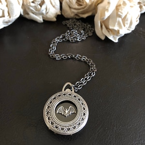 May include: A silver necklace with a round pendant featuring a black bat inside a decorative frame.