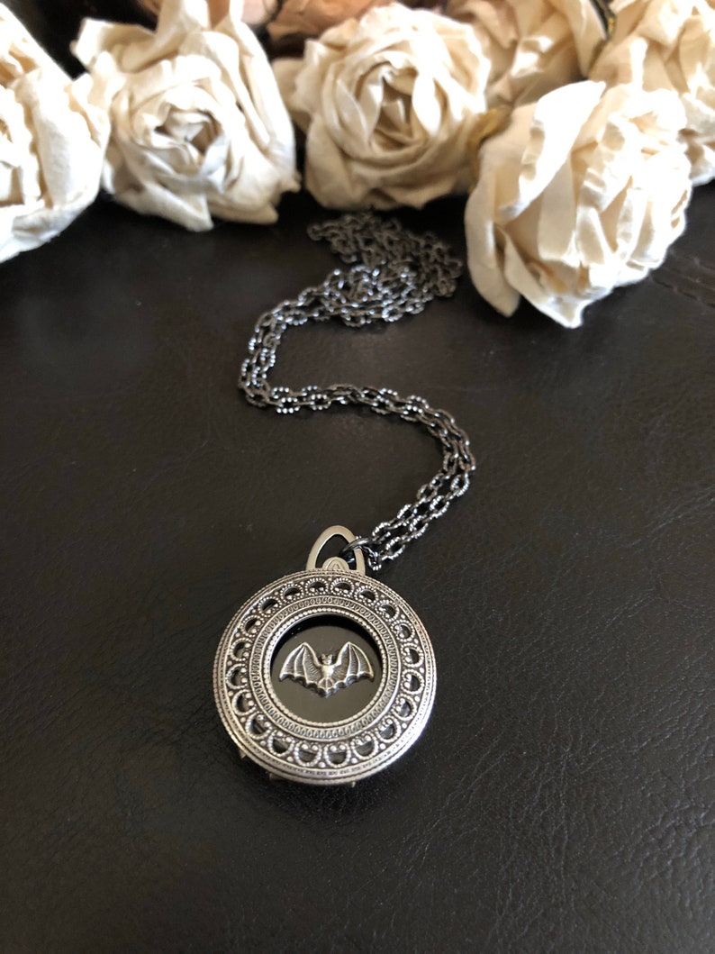 May include: A silver necklace with a round pendant featuring a black bat inside a decorative frame.