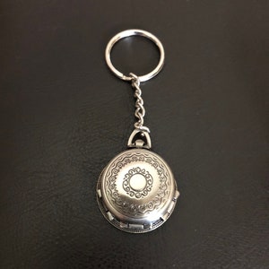 May include: A silver oval-shaped keychain with an intricate design and a small circle in the center. The keychain has a silver ring and chain.