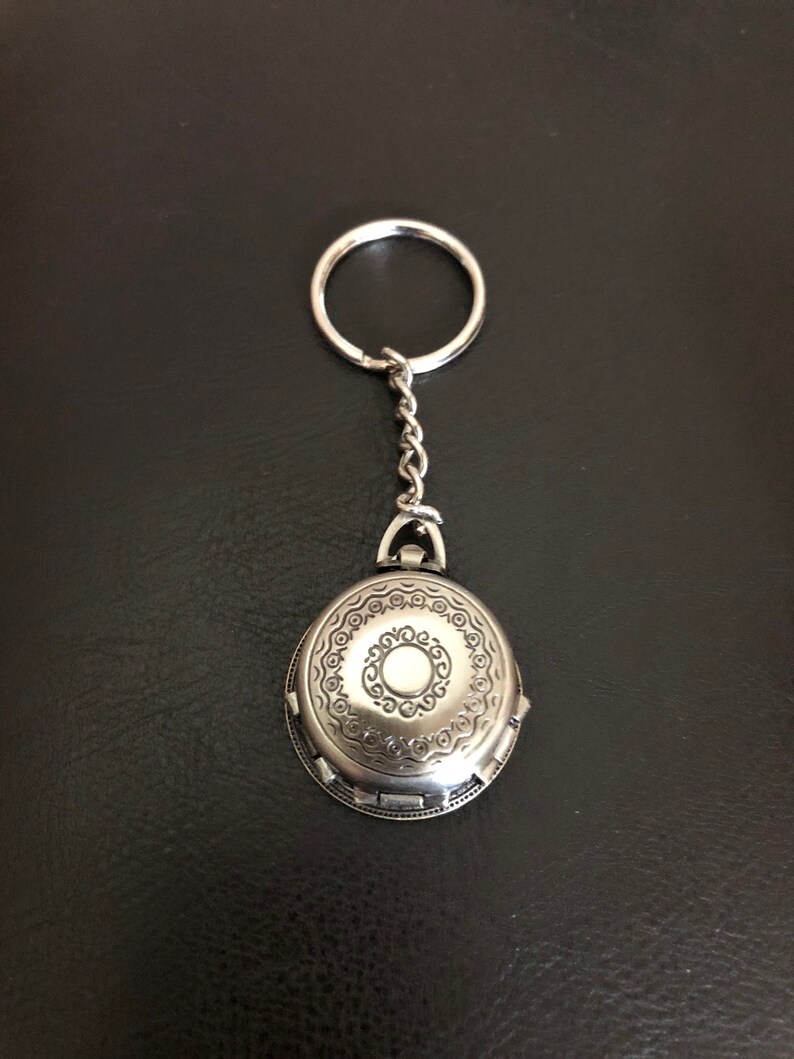May include: A silver oval-shaped keychain with an intricate design and a small circle in the center. The keychain has a silver ring and chain.