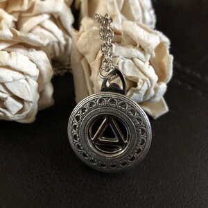 May include: Silver necklace with a round pendant featuring a black triangle inside a circle of decorative details.