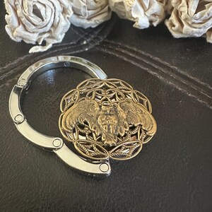 May include: A circular metal bag hanger featuring a bronze-toned owl emblem centered within an intricate filigree pattern. The hanger has a polished silver-colored folding hook mechanism that extends from the side of the decorative owl centerpiece.